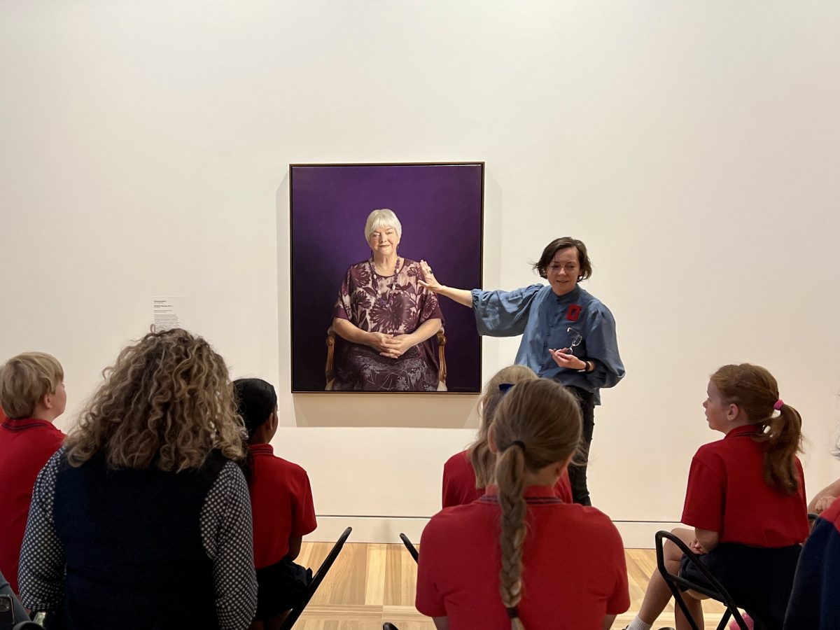 Woman gestures at portrait of Stephanie Alexander while school children sit and listen.