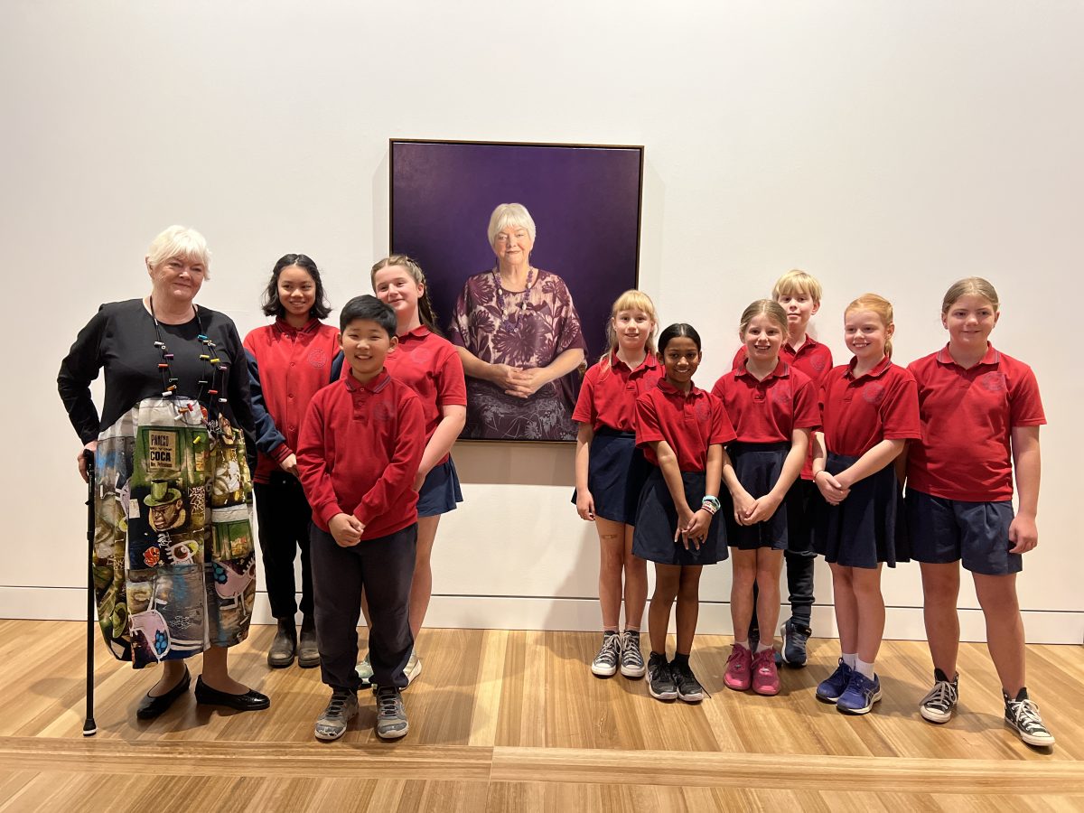 A group photo of school children in red uniforms in front of a portrait of Stephanie Alexander who stands alongside.