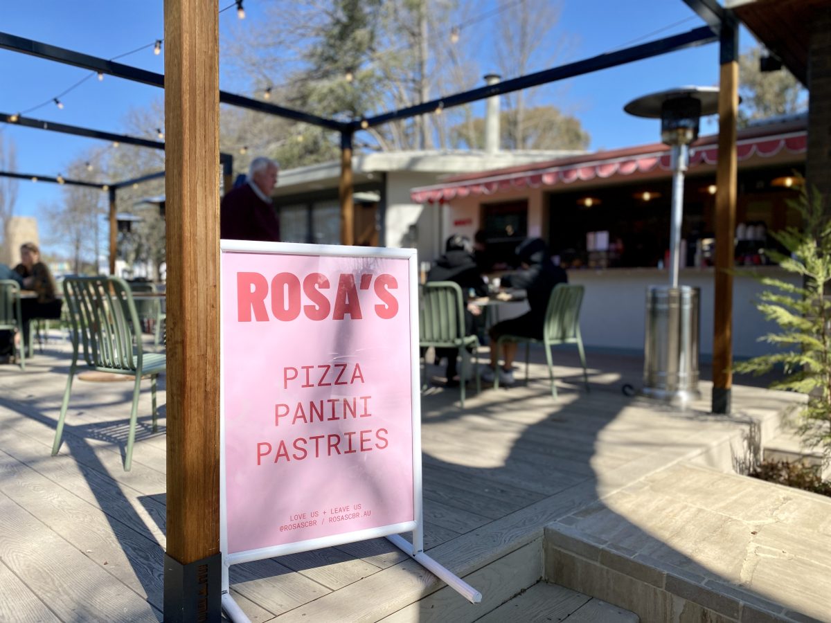 Pink sign with bold red text reading 'Rosa's Pizza Panini Pastries' sitting on a sunny deck.