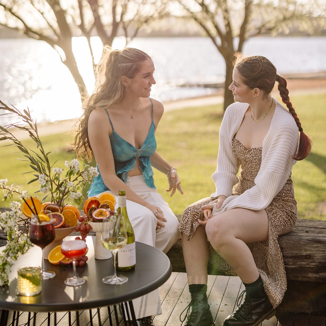 Two young women dressed in spring clothing sitting with cocktails by the lake.