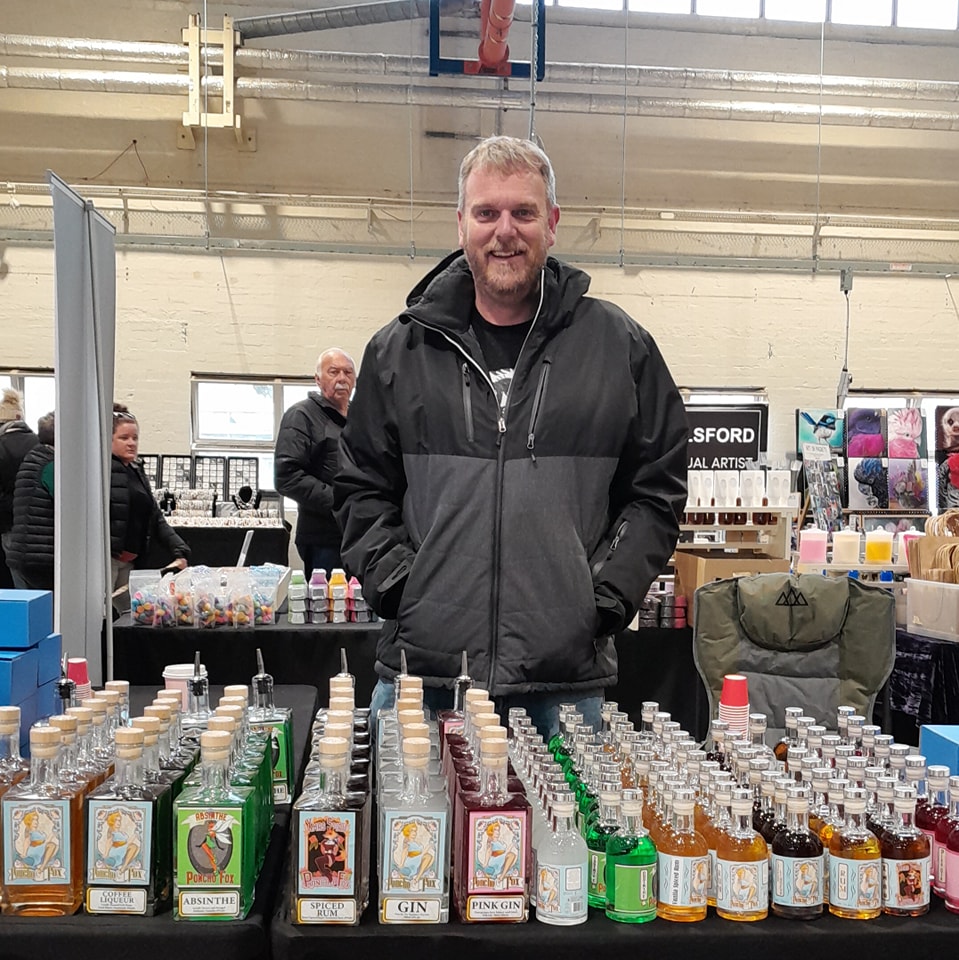 Patrick stands at a market stall with bottles of spirits.