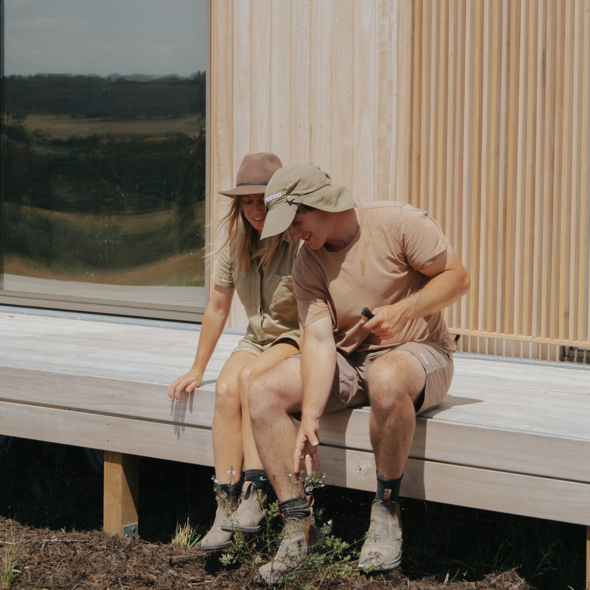 Two people wearing beige sit on the deck of a tiny house and look at the garden beds at their food. 