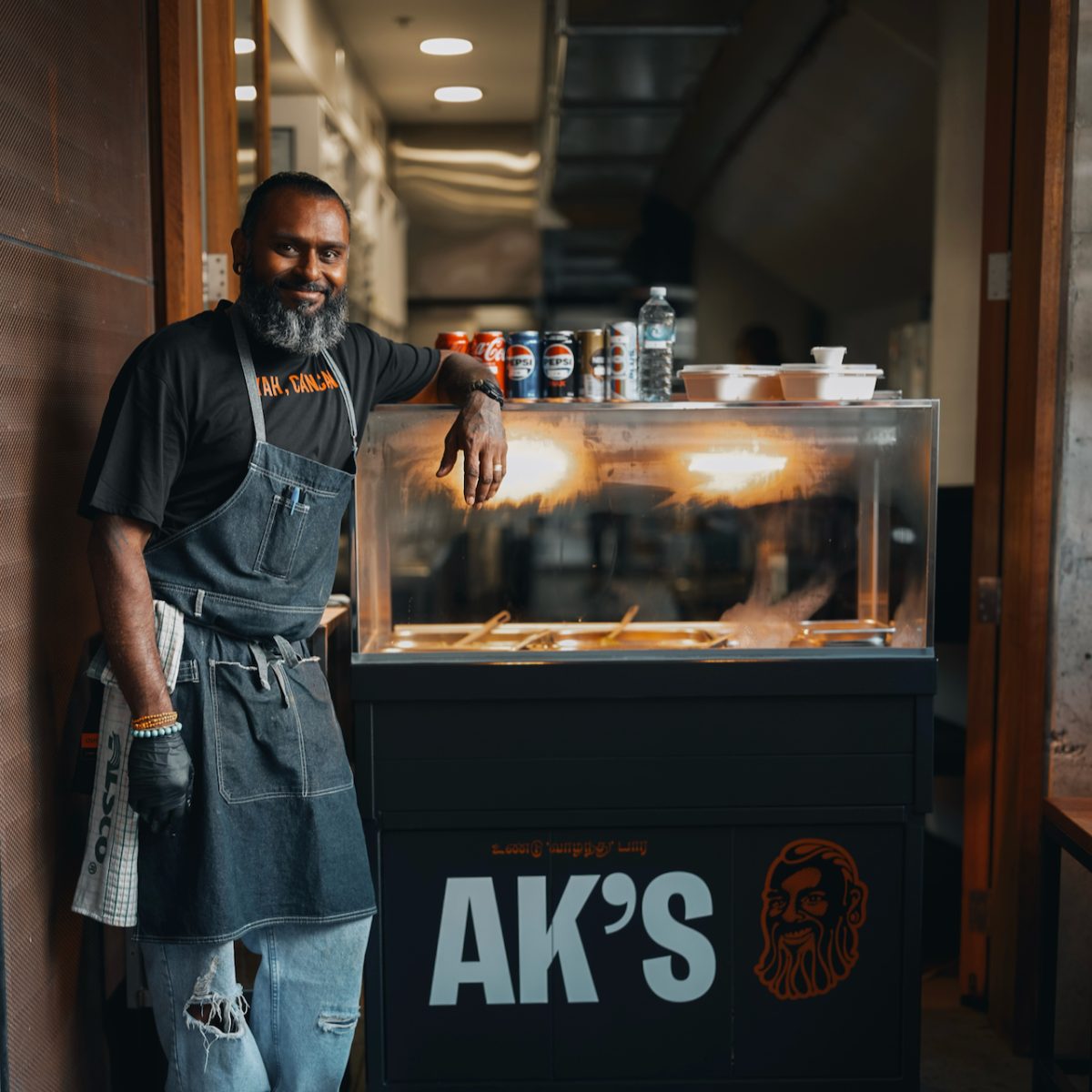 AK wears torn jeans, a black T-shirt and apron and stands next to a bain marie with a sign reading AK's.