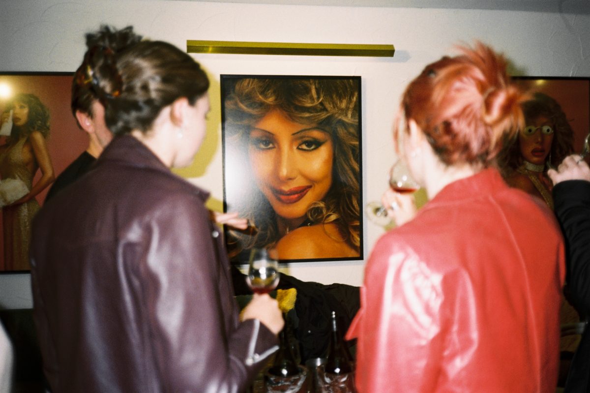 Two women sip wine in the foreground, and a large artwork of a woman's face is in focus in the centre. 