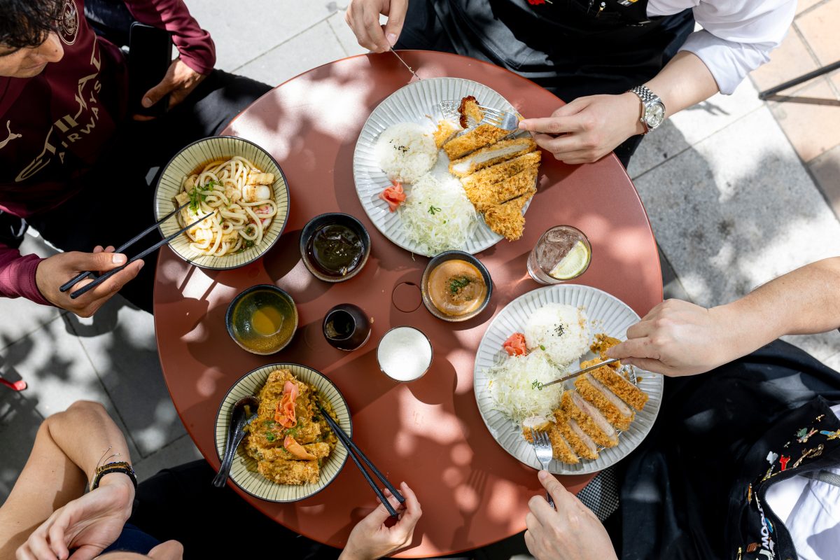 A top down shot of four people at a table in dappled sunlight with different dishes, drinks and sauces. 