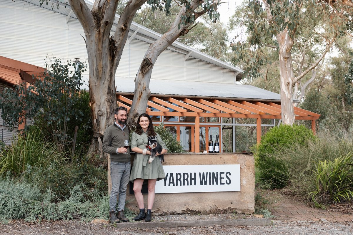 A man and woman stand together holding a dog in front of a sign reading Yarrh Wines. 