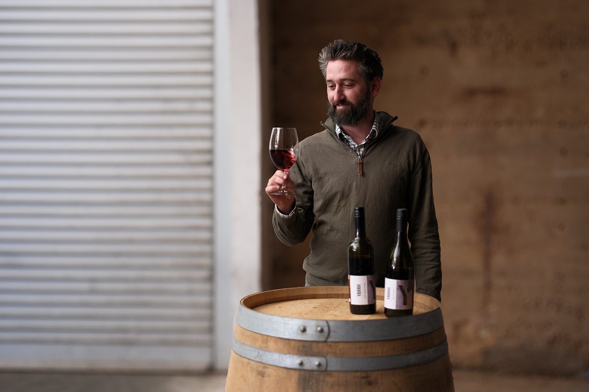 A bearded man examines a glass of red wine while standing next to an upturned wine barrel.