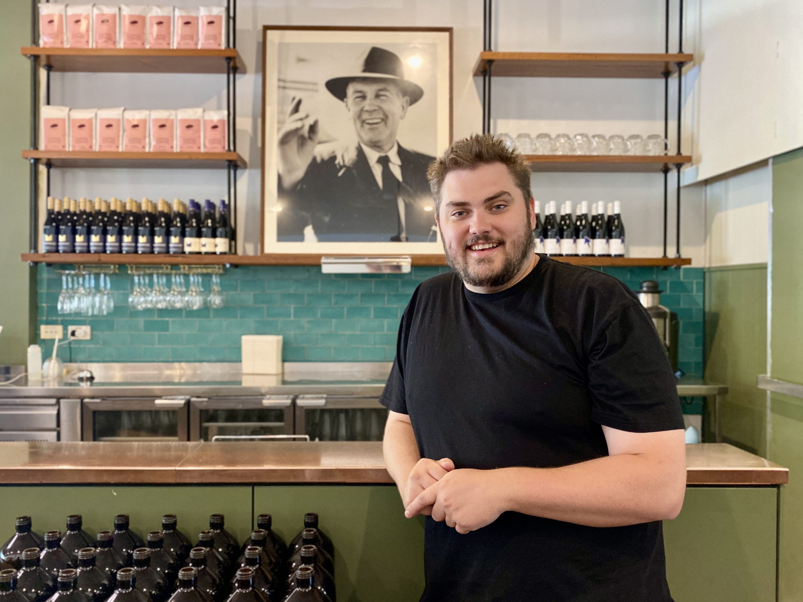 A man stands in front of a bar which features a black and white photo of Prime Minister Chifley.