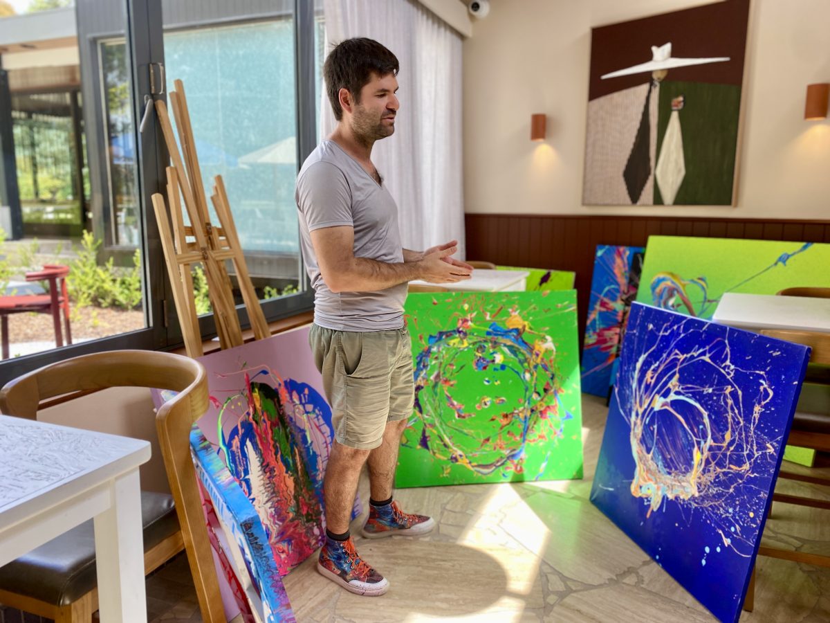 A man in shorts and T-shirt is surrounded by colourful canvases in a restaurant.