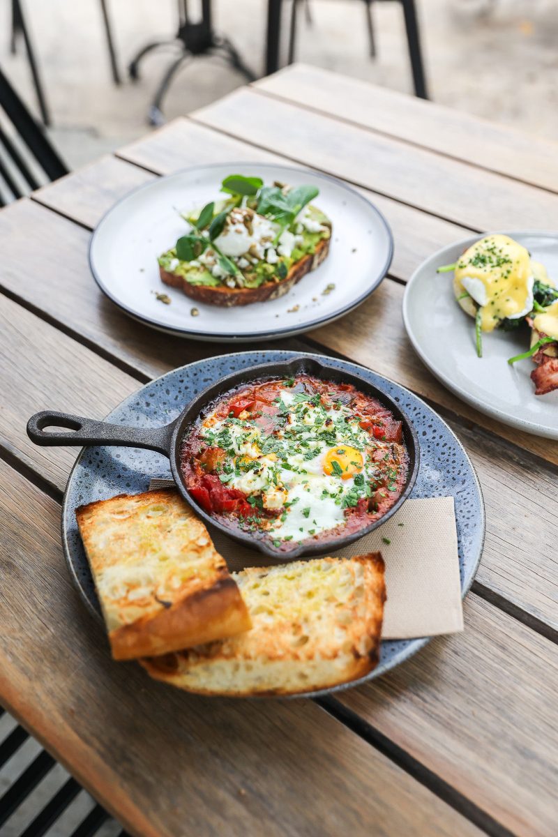 A plate with a small cast iron dish of eggs cooked in a red sauce, with chunky toast and other dishes in the background.