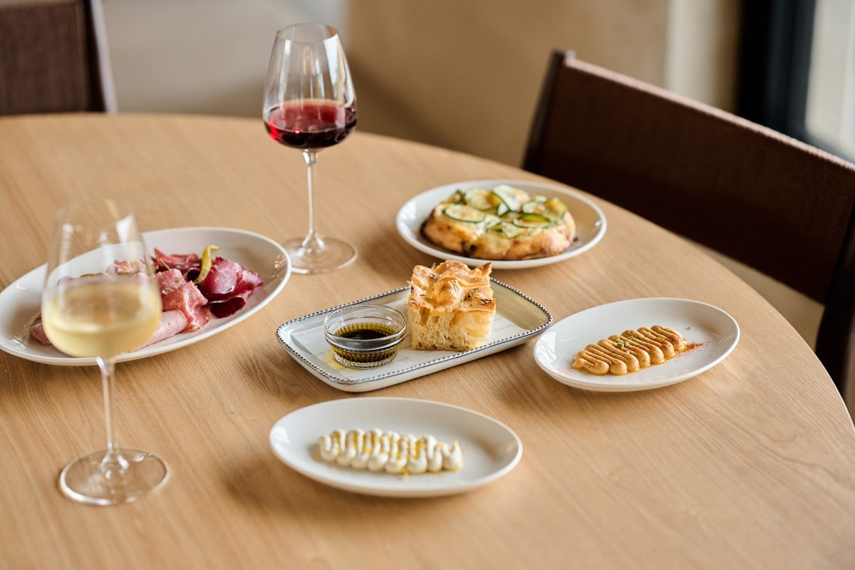 Several dishes on a table like dips, cured meats, flatbread and focaccia with glasses of wine.