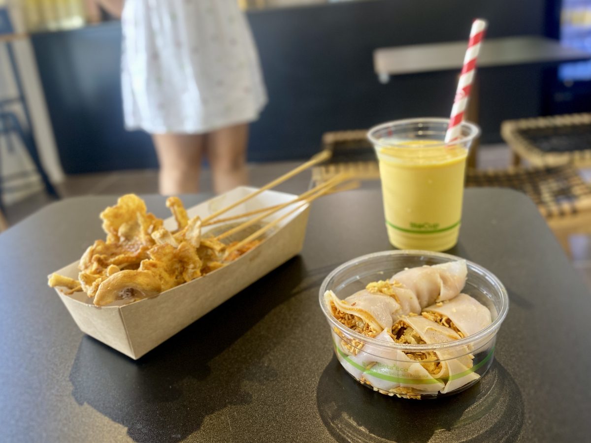 Takeaway dishes on a table with skewers, and a noodle dish with a yellow drink in the background.