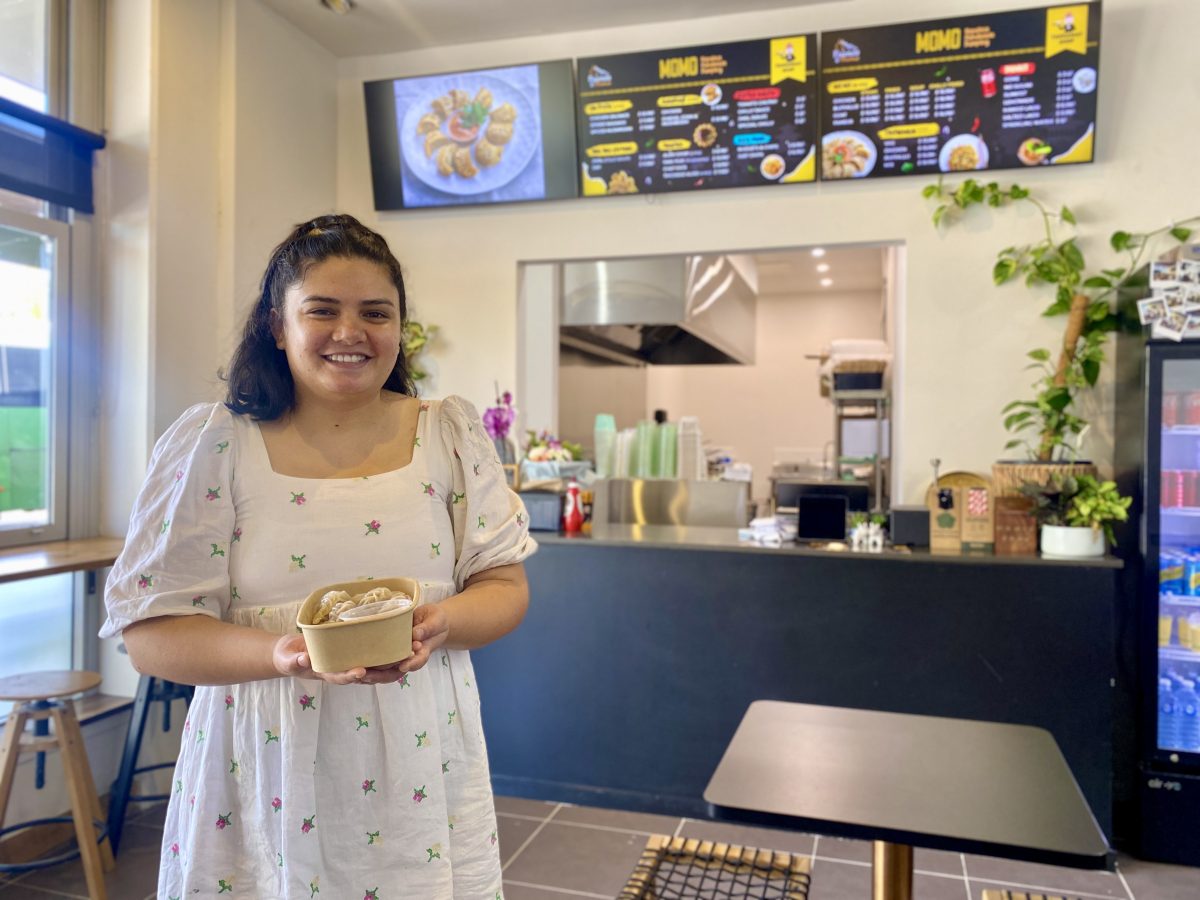 A woman in a white dress holds a box of momos in front of a cafe counter