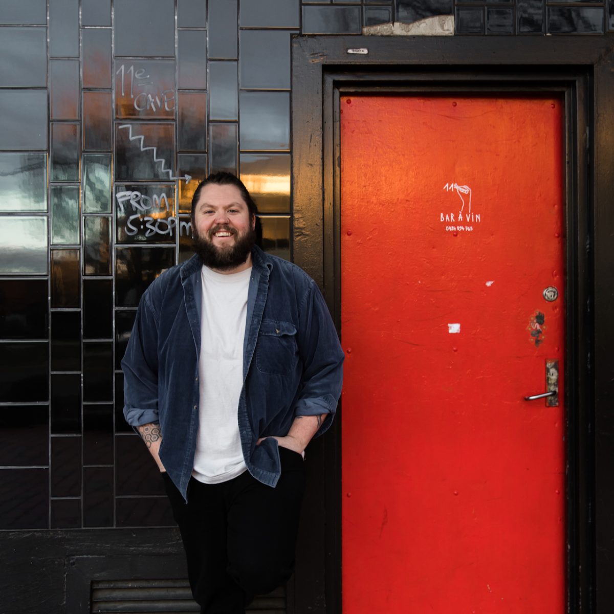 A bearded man stands next to a red door set in a black tiled wall.