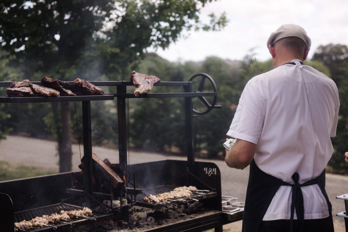 A man in a cap cooks meat on a charcoal BBQ.