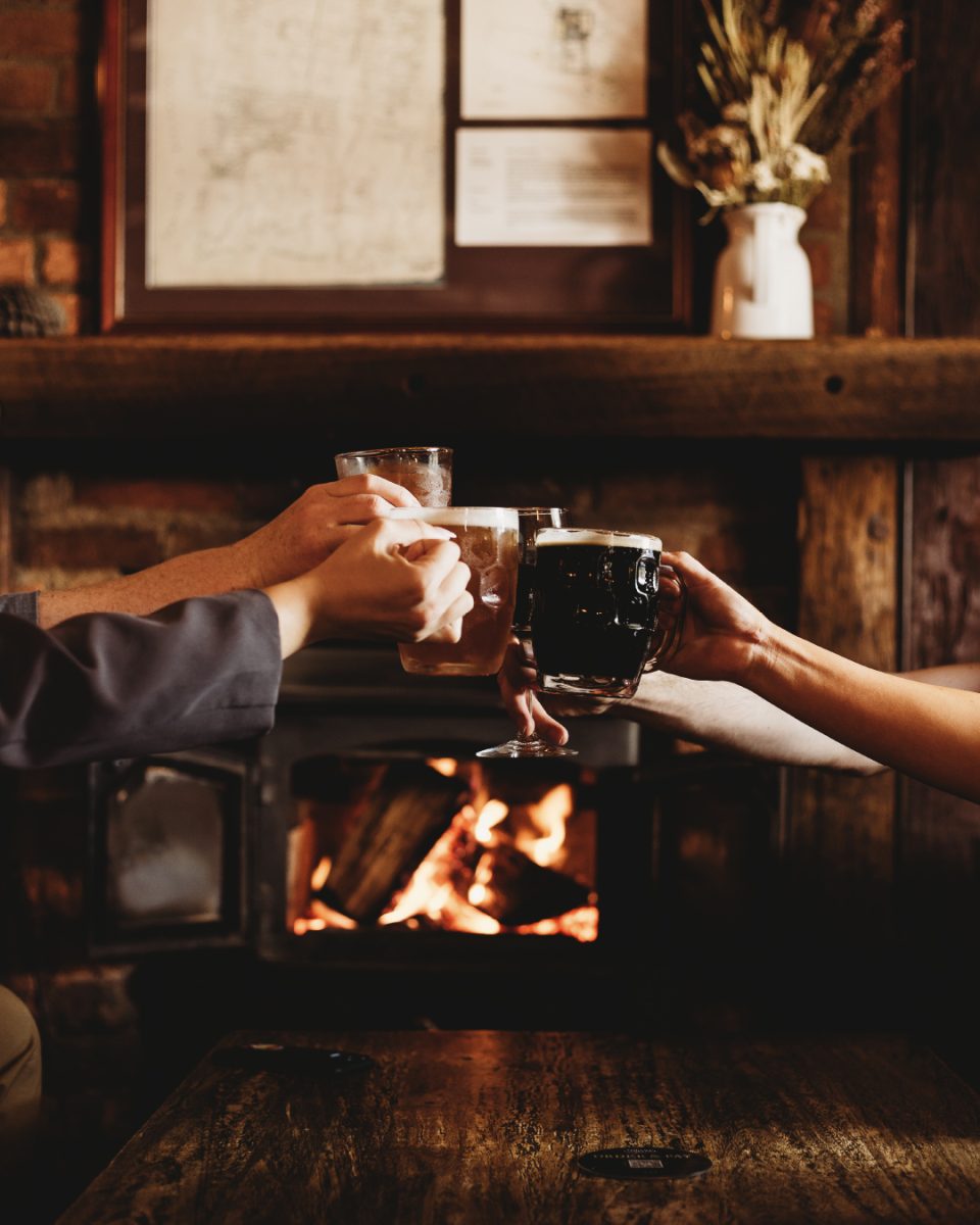 Four hands 'cheers' their pints of beers in front of a roaring fireplace.