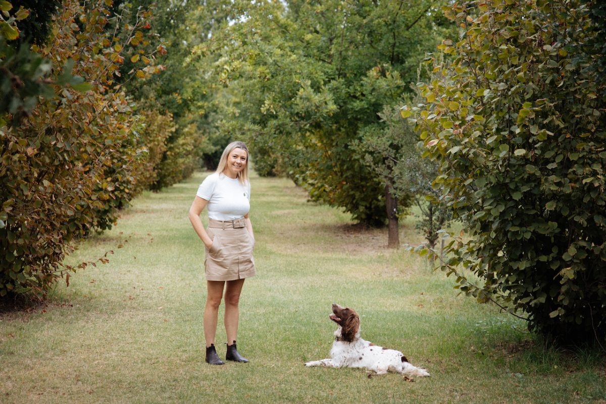 A blonde women stands on grass surrounded by trees with a dog at her feet