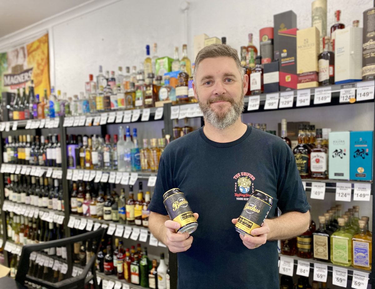 A man holds two cans of beer and stands in front of a shelf of alcohol bottles.