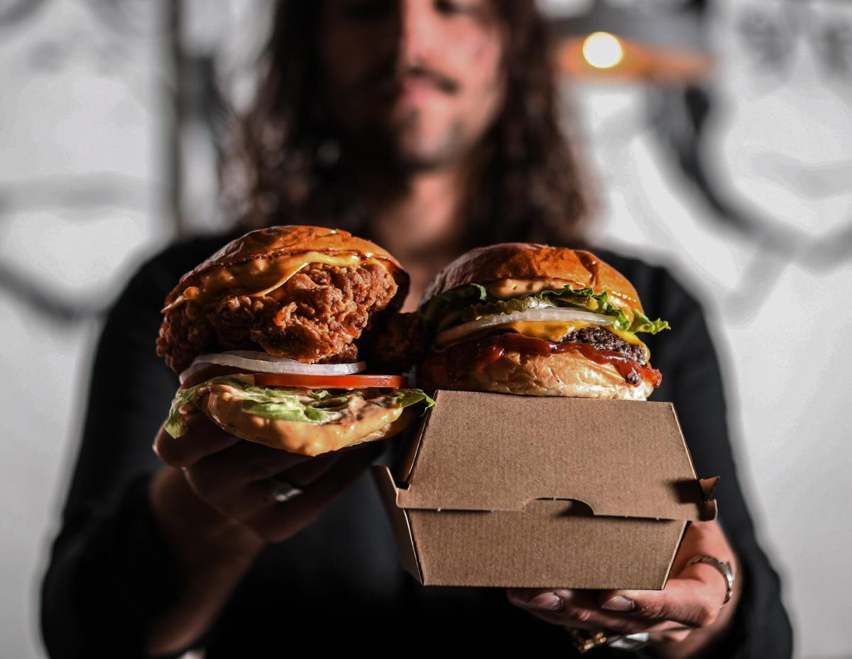 A bearded man holds a stack of burgers in both hands.
