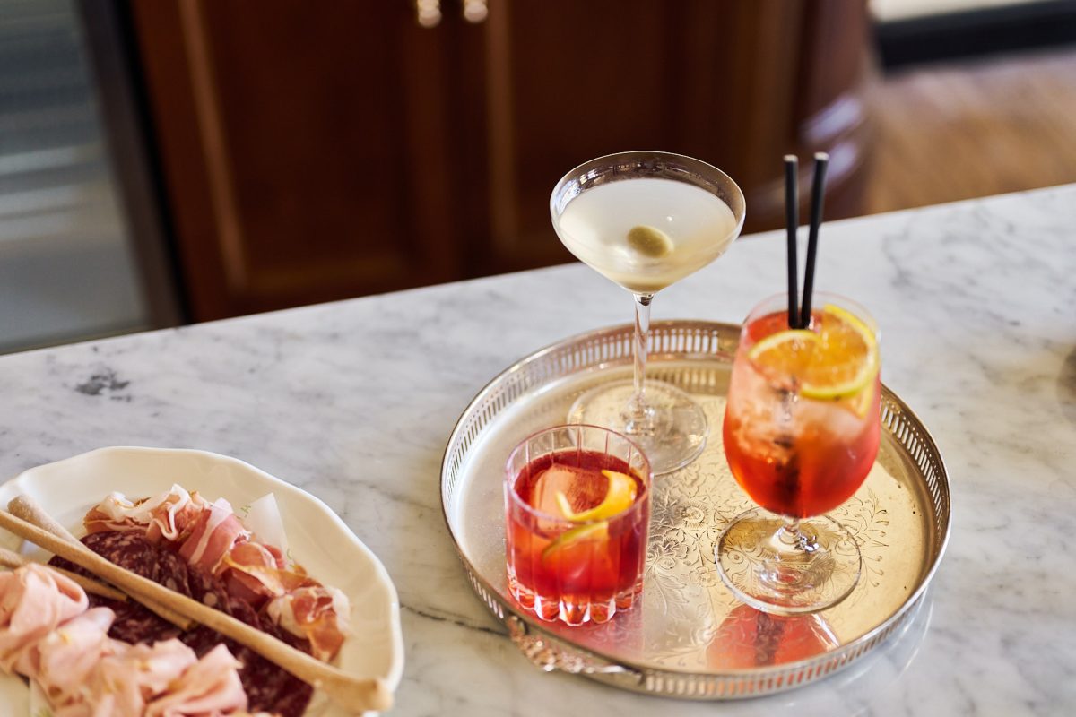 Three cocktails on a silver tray next to a plate of salami on a marble table.