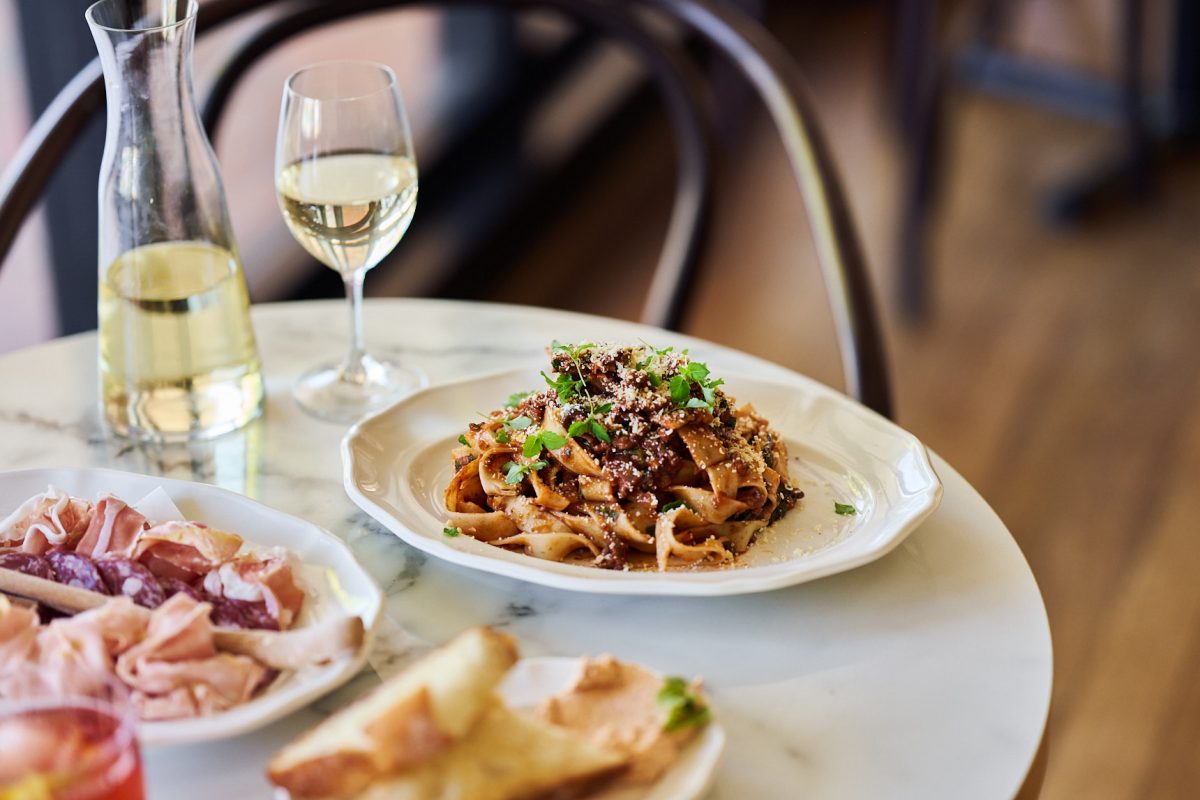 A plate of pasta with a meat ragu next to a glass of white wine on a marble table.