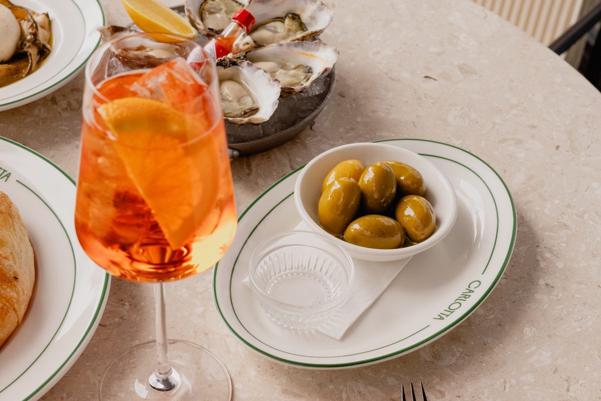 A glass of aperol spritz and six large sicilian olives in a dish,
