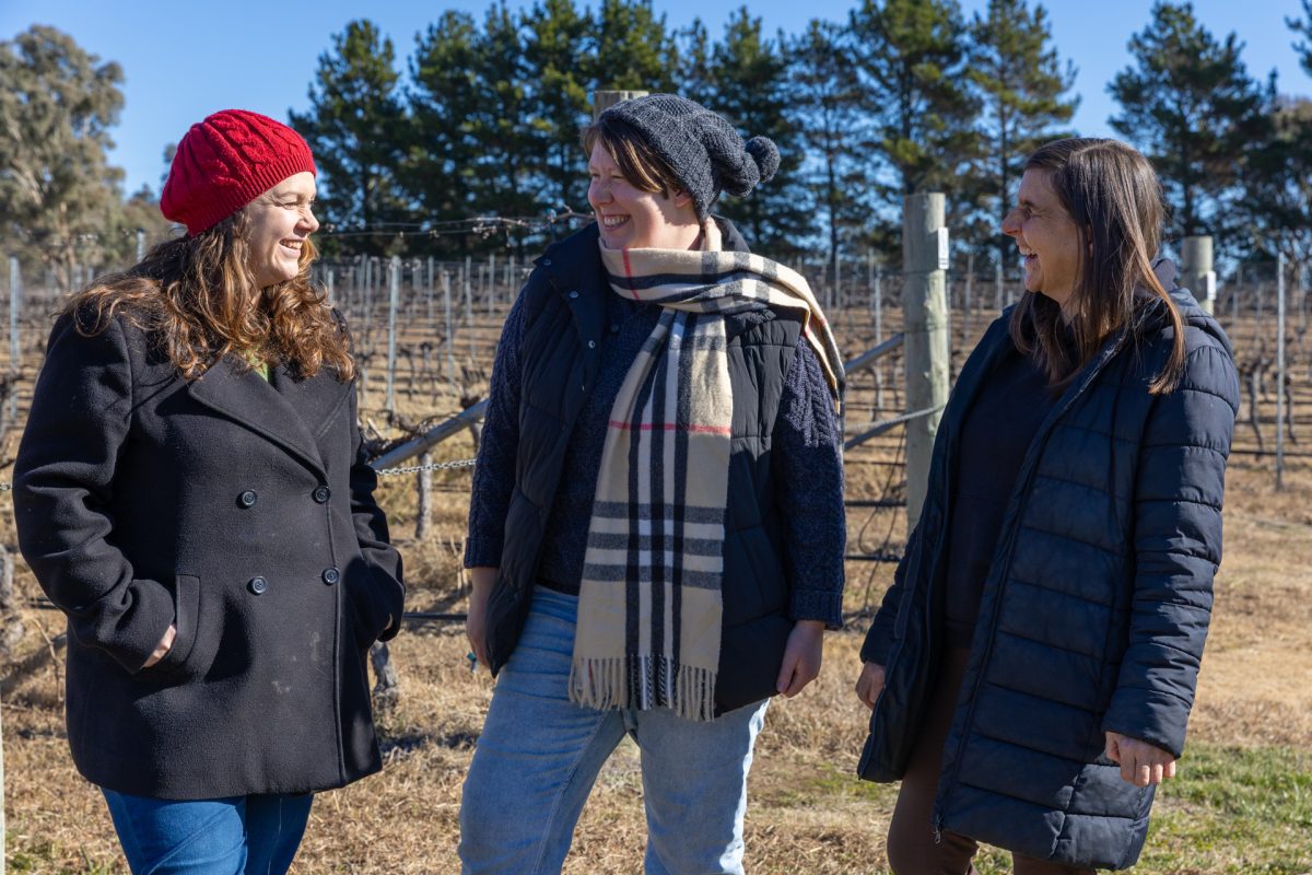 Three women rugged up in warm clothes stand smiling casually in a vineyard.