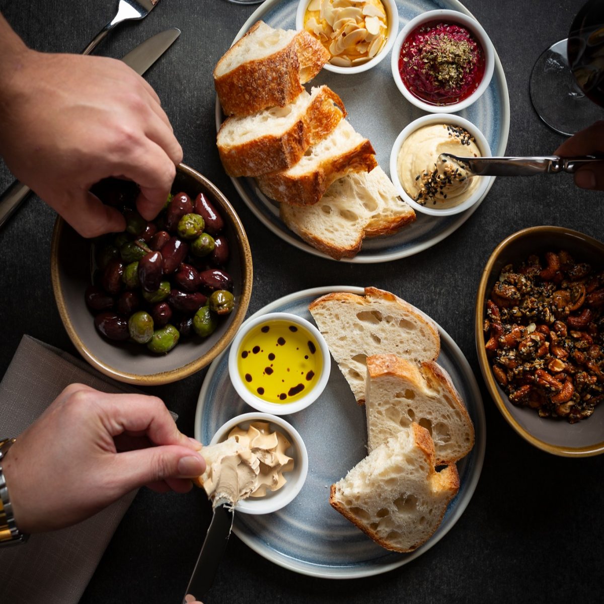 A table cluttered with plates of bread, olives and dips. Hands are reaching to take food.