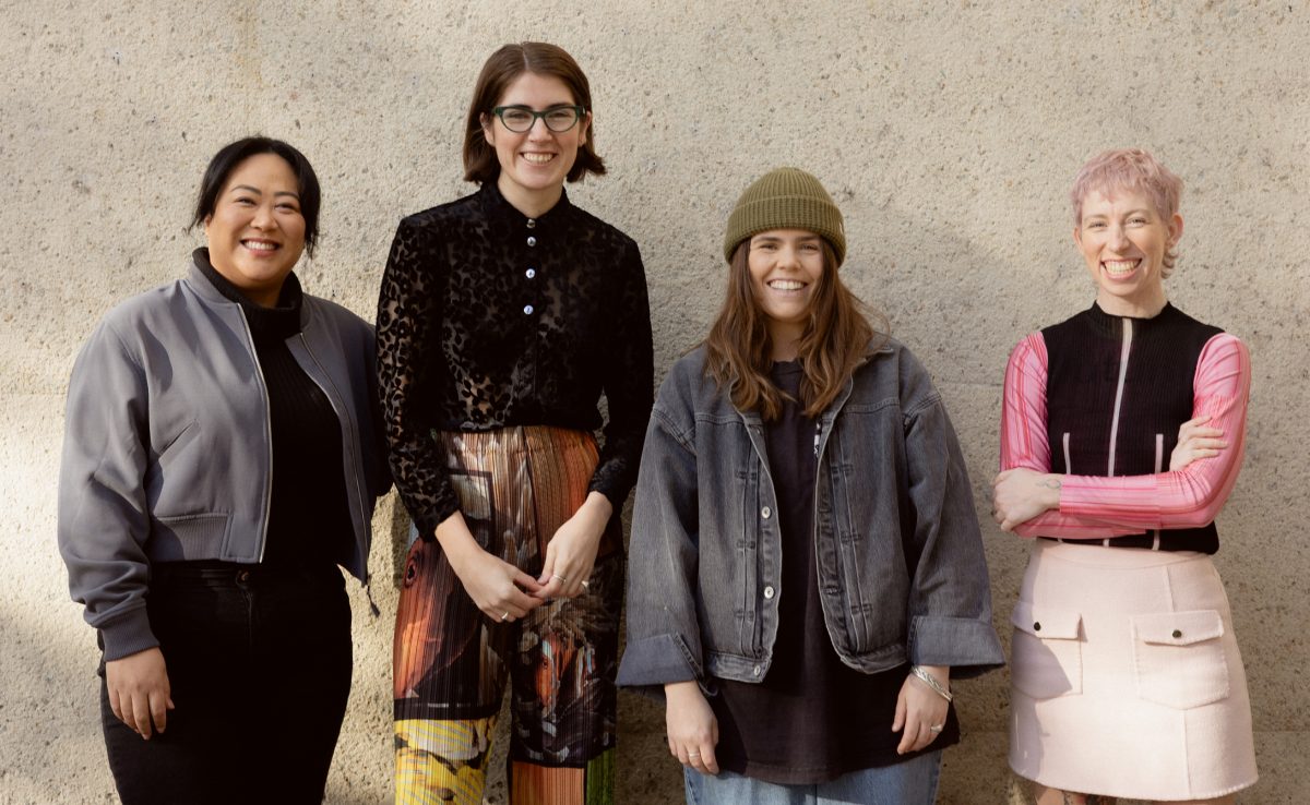 Four stand in front of a textured concrete wall. They are standing casually and smiling at the camera.