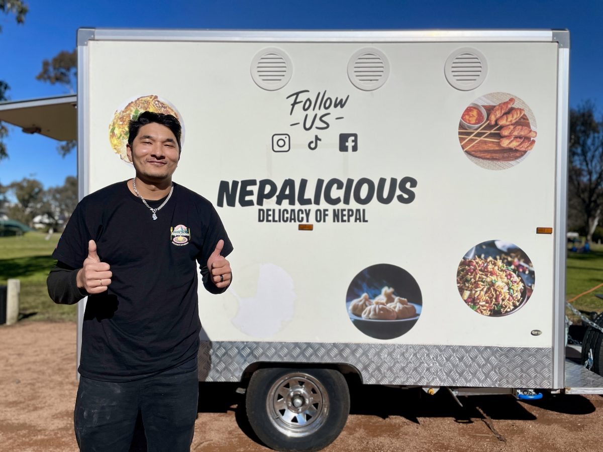 A Nepalese man wearing black gives two thumbs up to the camera, standing in front of a food truck which reads "Nepalicious, delicacy of Nepal"
