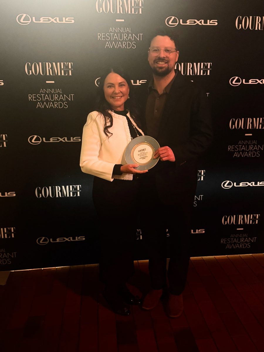 A woman and taller man hold a trophy plate in front of a background with the Gourmet Traveller logo
