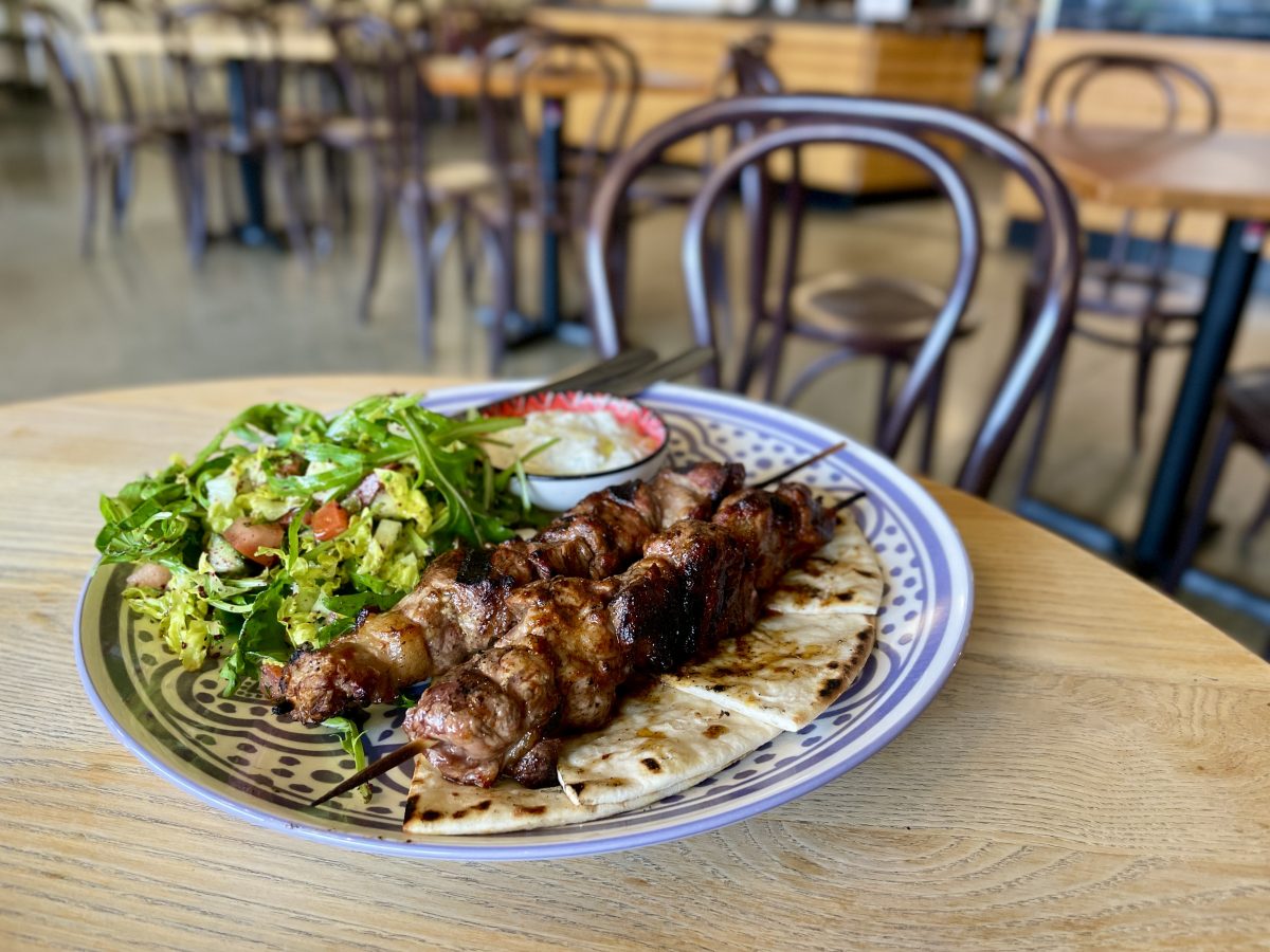 A blue plate with two delicious looking lamb skewers on triangles of pita bread, with a salad and ramekin of yogurt sauce.