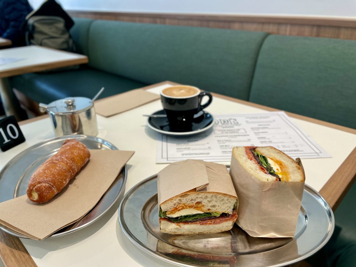 A doughnut filled with custard, a coffee and a good looking sandwich wrapped in brown paper.