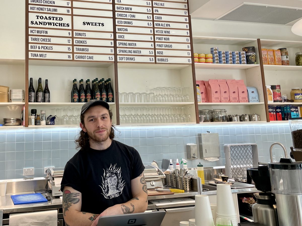 Young man in cap and black shirt stands behind cafe counter. 