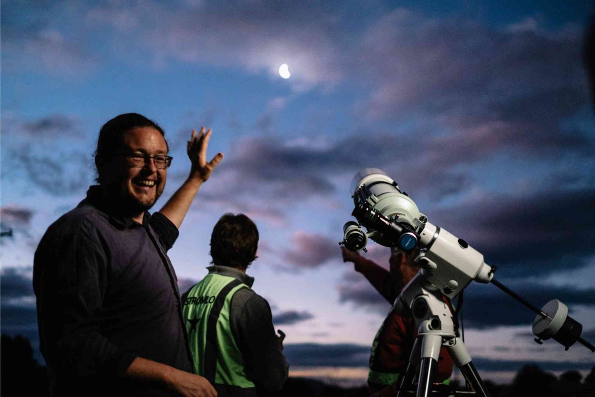 Astrophysicist Dr. Brad Tucker stands beside a telescope, gesturing to the moon in the background.