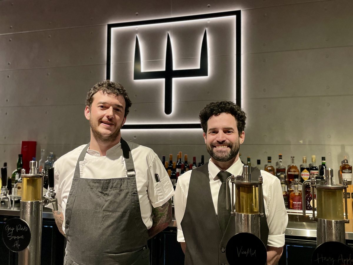 Two men - one in chefs jacket, one in shirt and waistcoat - stand in front of VICE's trident logo. 