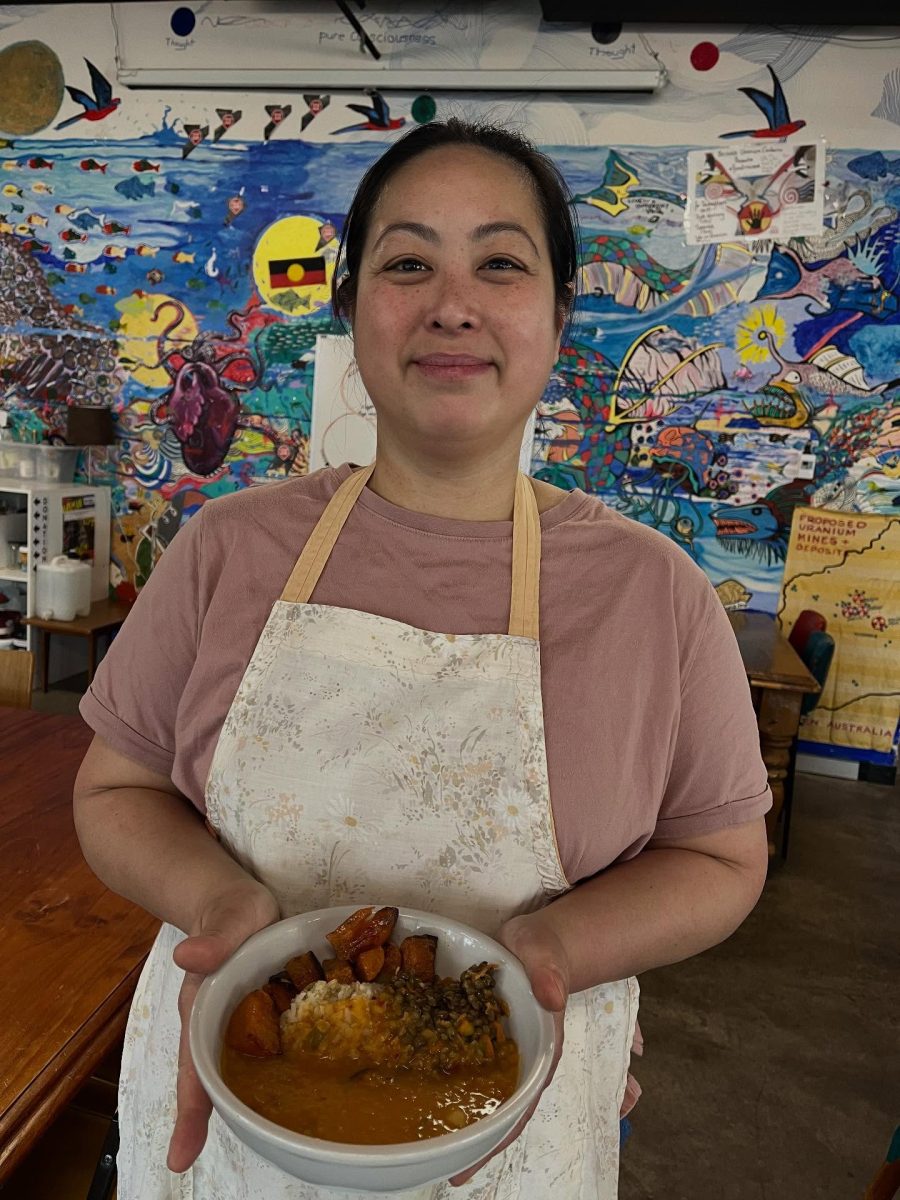 A Singaporean woman in a pink T-shirt holds a bowl of food in front of a colourful mural.