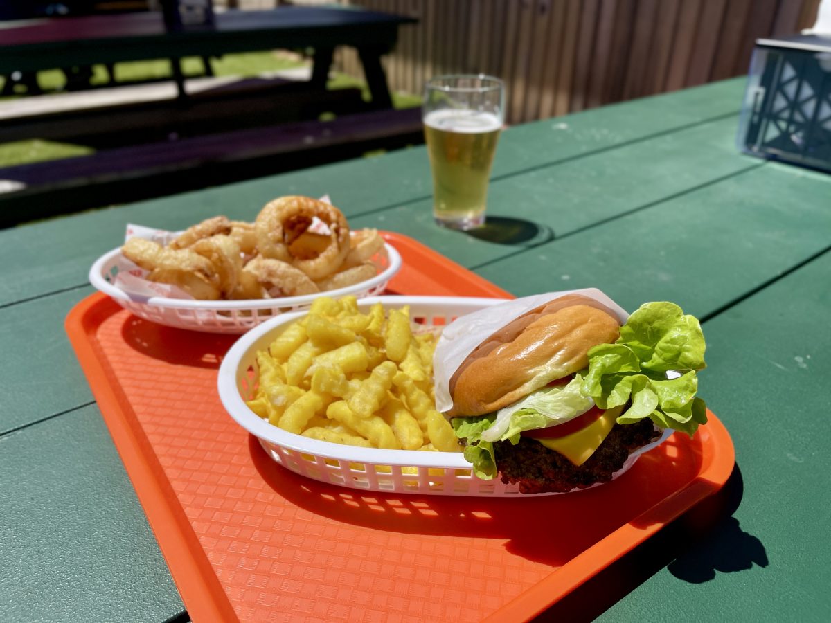Two plastic dishes on a red tray in the sun: one containing a burger and crinkle cut fries, the other with onion rings. Behind, a beer.