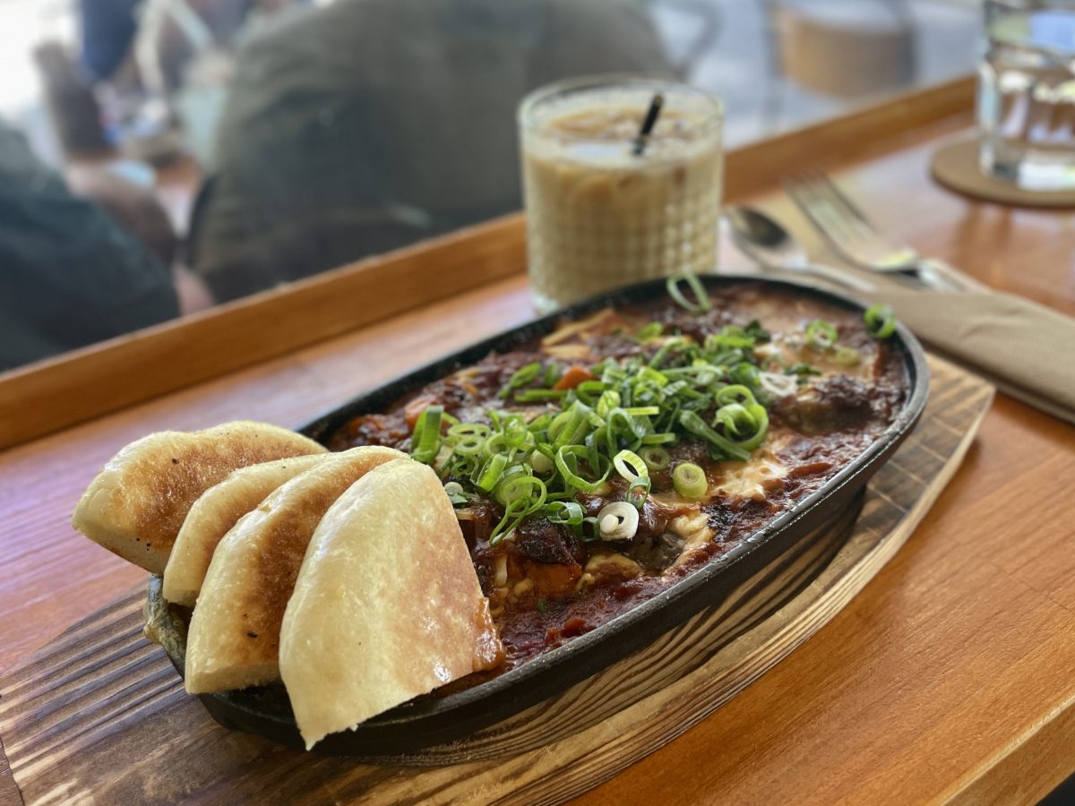 A cast iron dish on wooden board with a tomato sauce, scattered with spring onion and served with triangles of bread.
