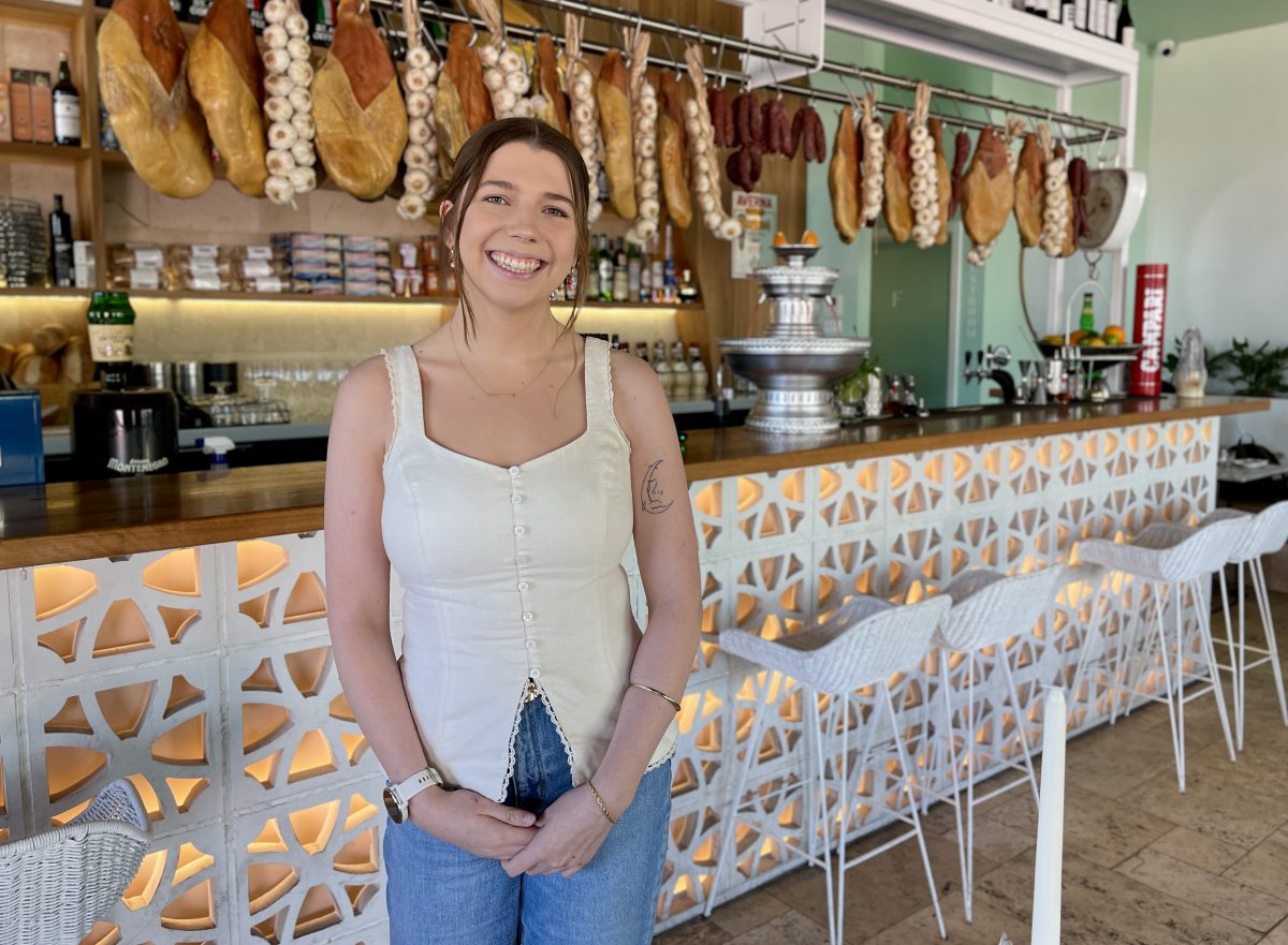 A young woman in white top stands in front of a bar hung with hams and garlic braids.