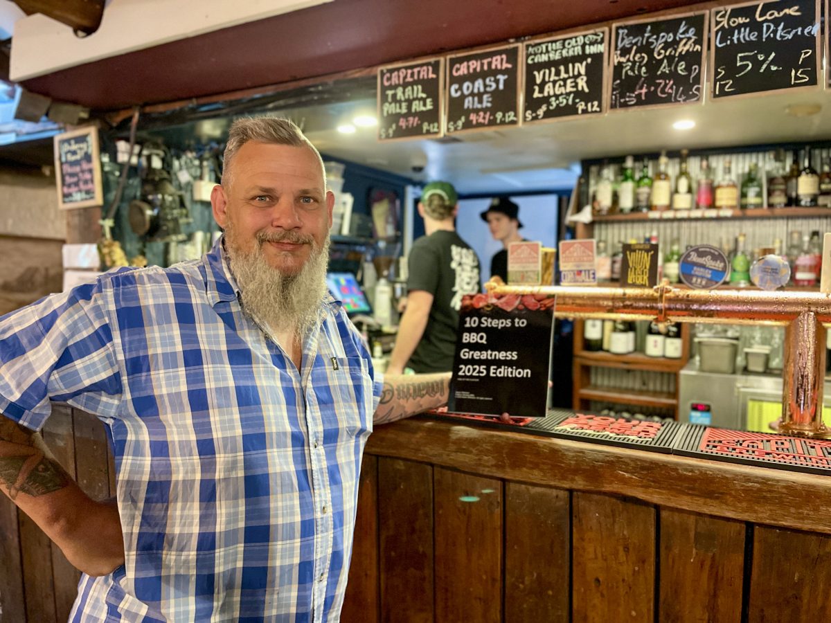 A bearded man in blue checked shirt holds a copy of "Ten steps to Barbecue Greatness" and leans agains a bar.