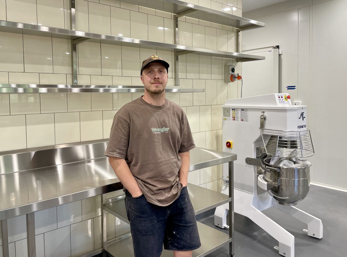 Lachlan leans against a shiny stainless steel bench. In the background, a large commercial mixer.