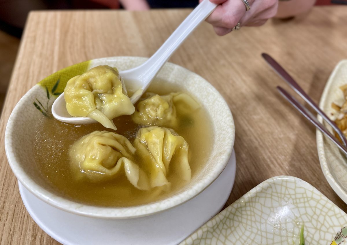 A white hand with silver rings lifts a flat, white Chinese-style soup spoon with a plump dumpling from a bowl of broth. 