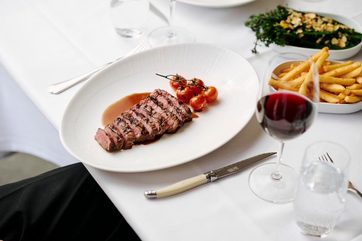 A plate with sliced steak and cherry tomatoes. A glass of red wine and a plate of chips is also on the white table