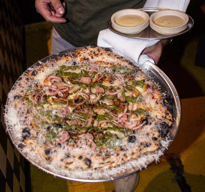 A server holds a tasty looking pizza on a metal tray, and also holds two ramekins of sauce.