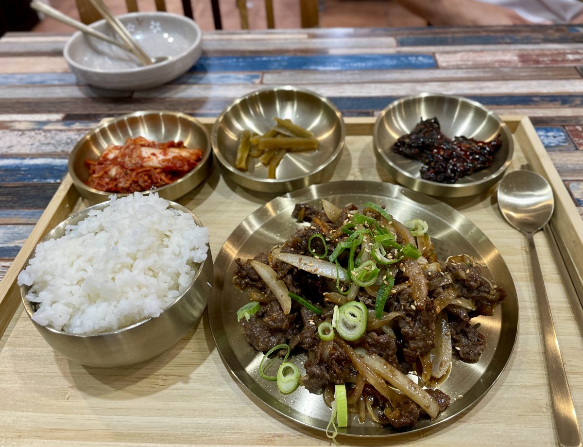 A wooden tray with shiny brass-looking bowls filled with rice, pickles and kimchi and a larger dish of stir fried beef.