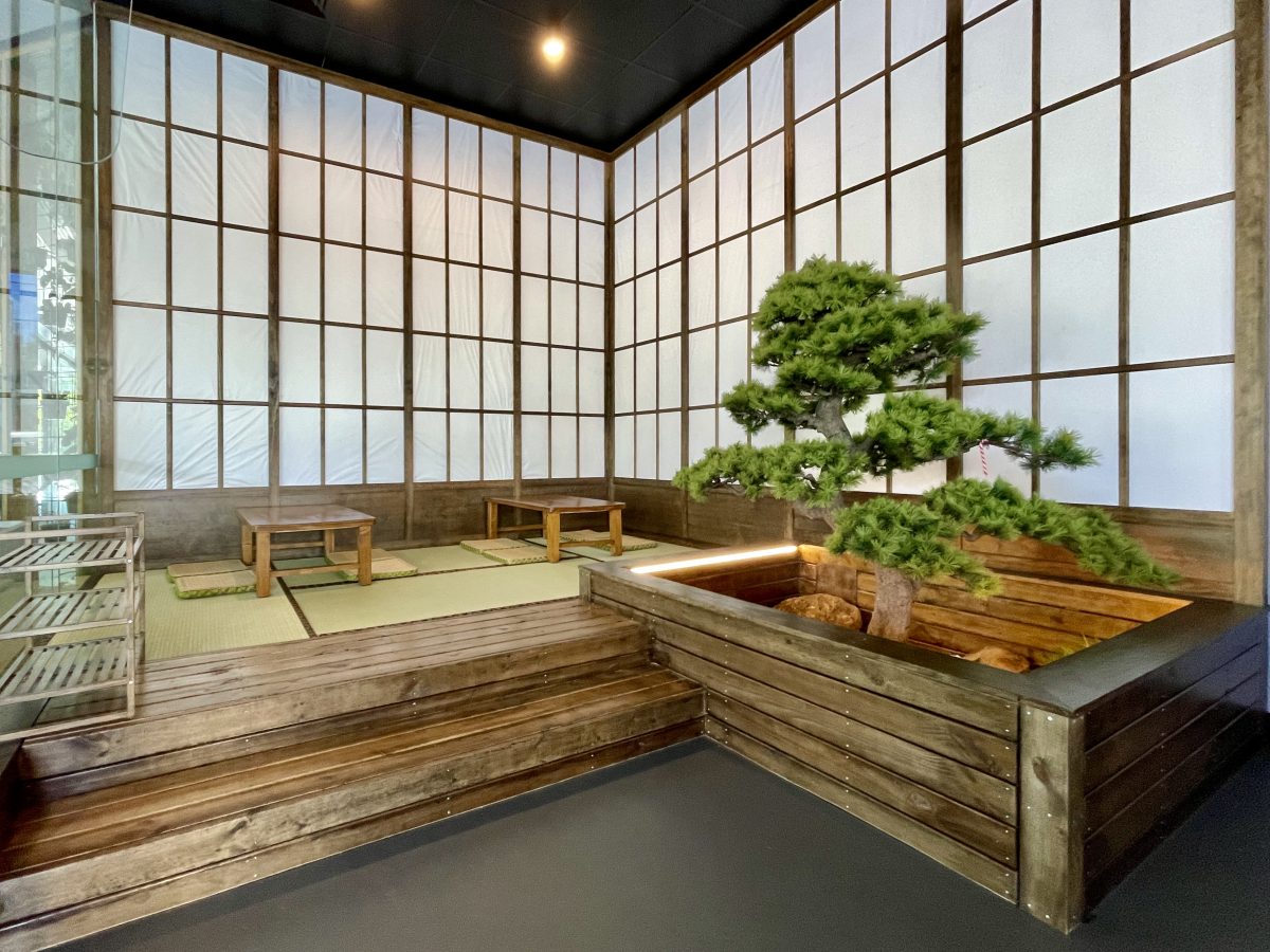 A corner of a cafe decorated in a traditional Japanese style with low tables, woven mats and paper screens on the walls. There is also an impressive Bonsai in a wooden planter box.