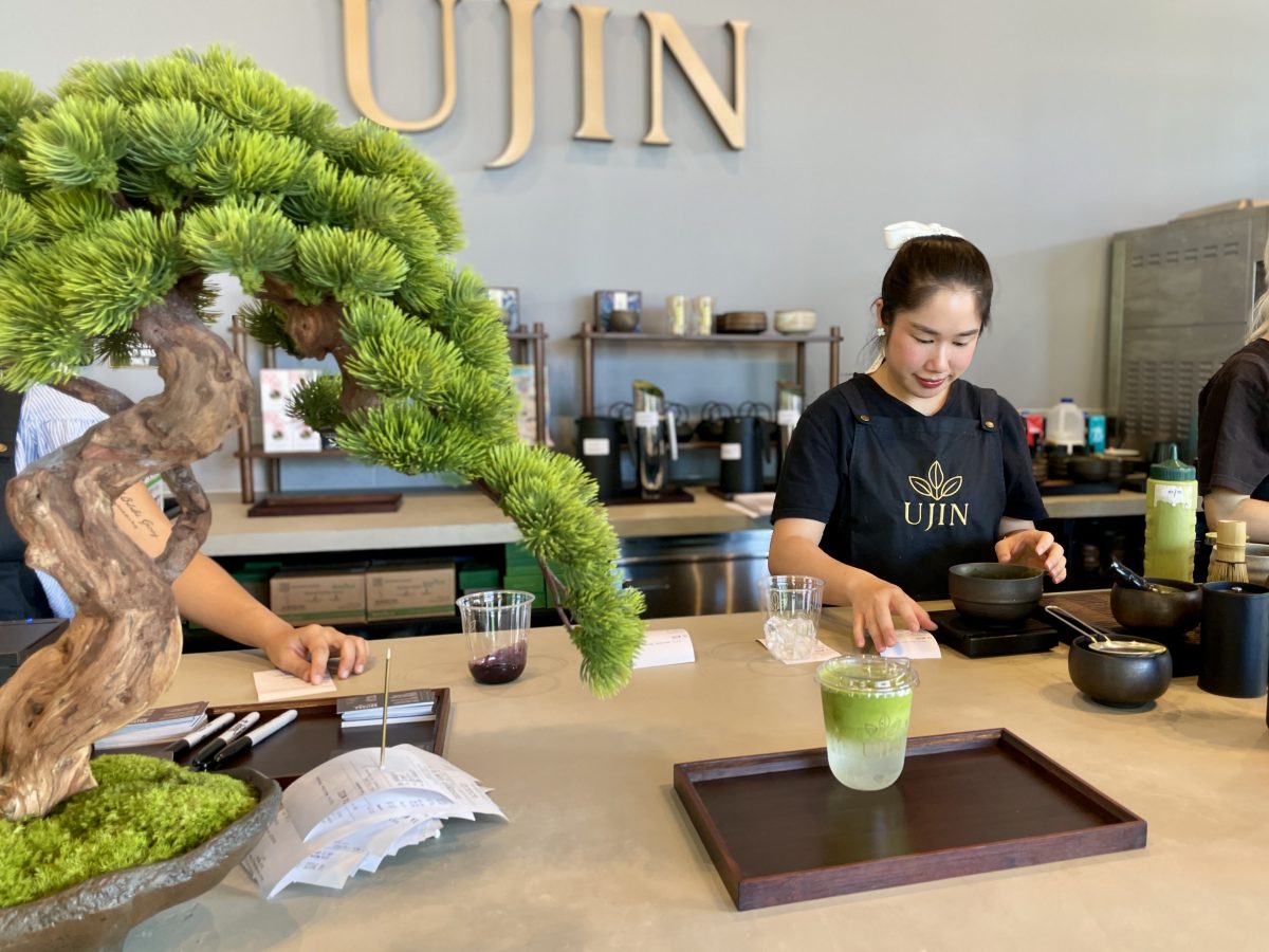 A bonsai tree on a cafe counter next to a green drink in a clear takeaway cup. A woman in a black apron reading 'Ujin' works in the background.