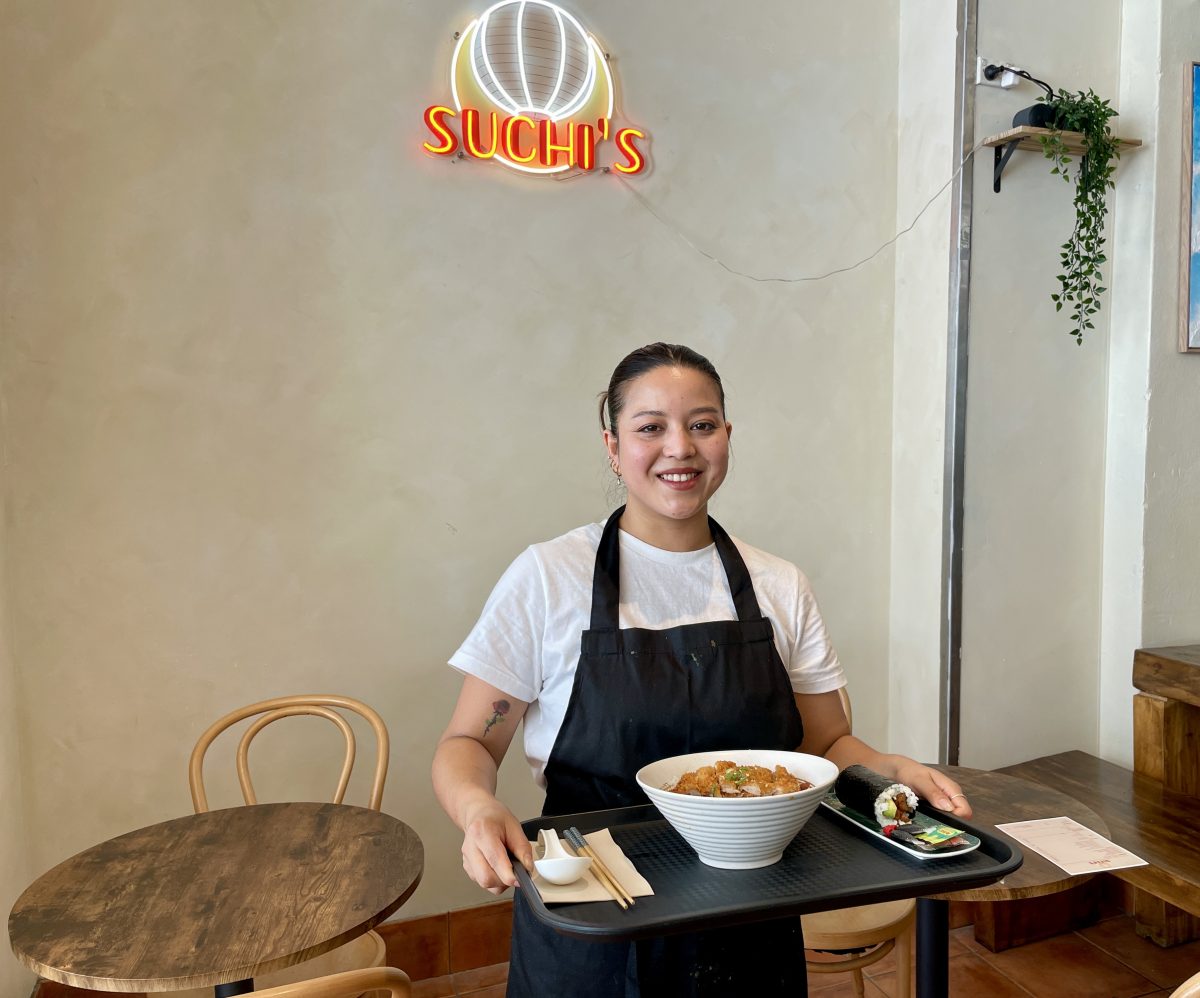 A woman in white tshirt and black apron holds a tray of food in front of a neon sign reading 'Suchi's'.