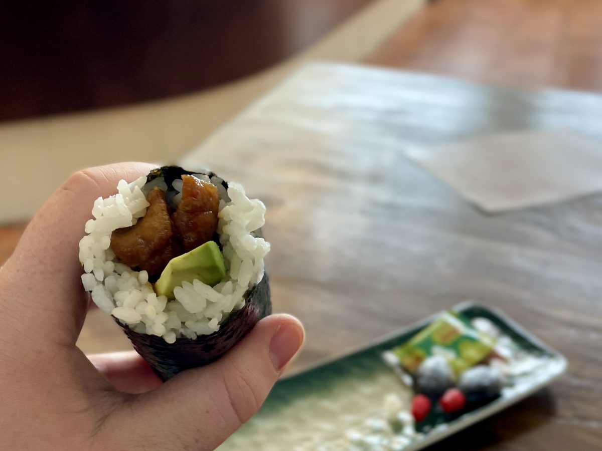 A white hand holds a sushi hand roll filled with chicken and avocado